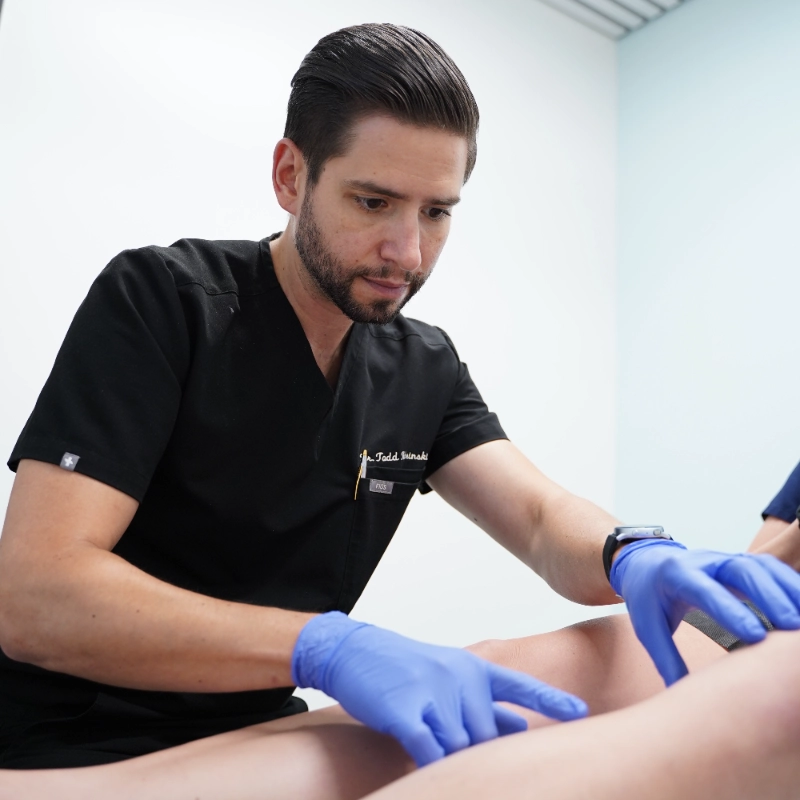 Dr. Todd Kobrinski examines a patient's leg during a vein assessment in a clinical setting. Wearing blue medical gloves, the provider performs a physical examination of the lower extremity in a bright, clean medical office. This evaluation is part of a comprehensive vein treatment consultation to assess for conditions such as varicose veins or venous reflux.